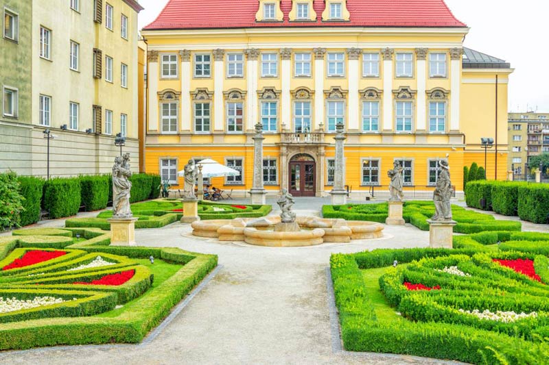View Of The Royal Palace – History Museum Baroque Style Building And Beautiful Garden With Sculptures In A Courtyard.