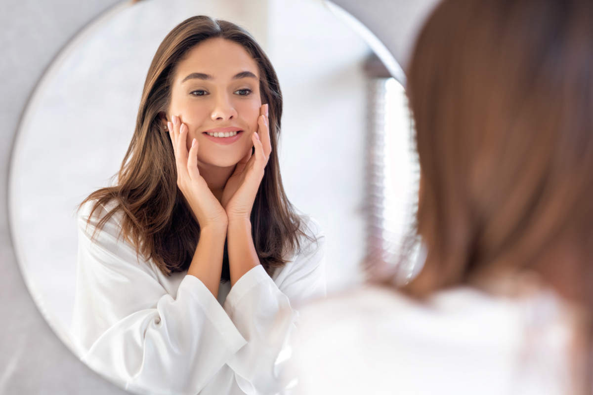 Skincare Concept. Pretty Young Woman Touching Her Face While Looking At Mirror