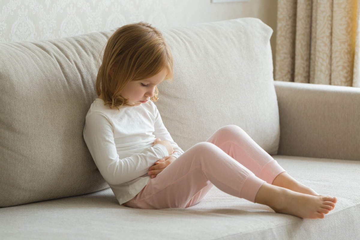 Little Girl Sitting On Sofa And Touching Painful Belly With Arms At Living Room. Child Suffering From Stomach Ache At Home.