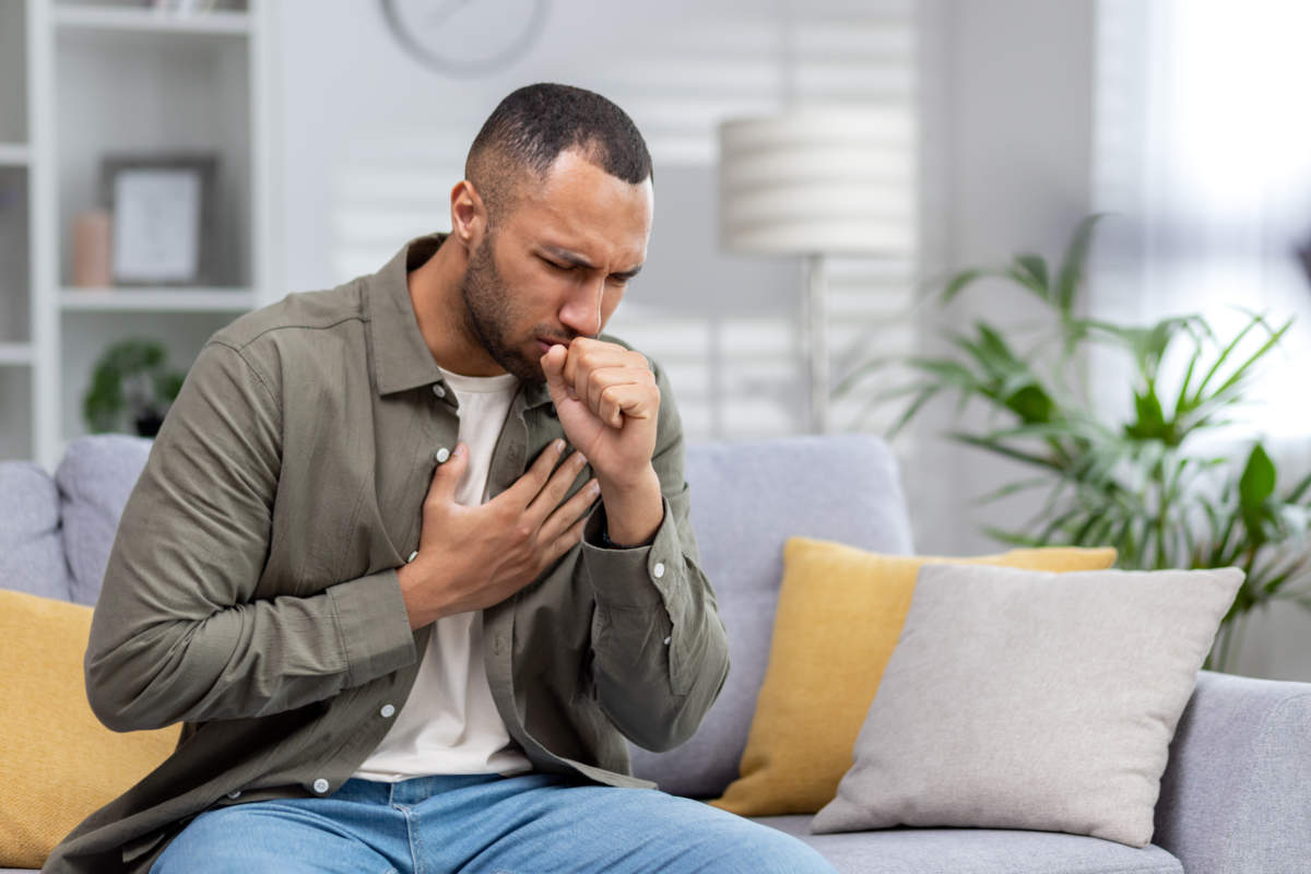 Young African American Man Sitting On The Couch At Home And Coughing. Suffers From An Attack Of Asthma, Allergies. He Holds His Chest, Covers His Mouth With His Hand