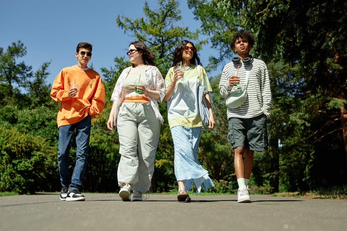 Four Teenagers Walking Along Park Path Chatting Enjoying Drinks Outdoors