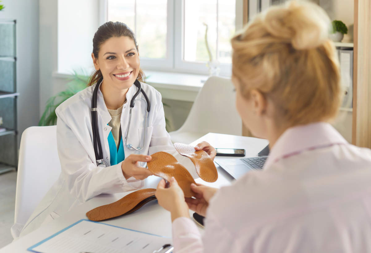 Orthopedist Offering Orthopedic Insoles To Patient At Appointment, Holding Two Orthotic Insets
