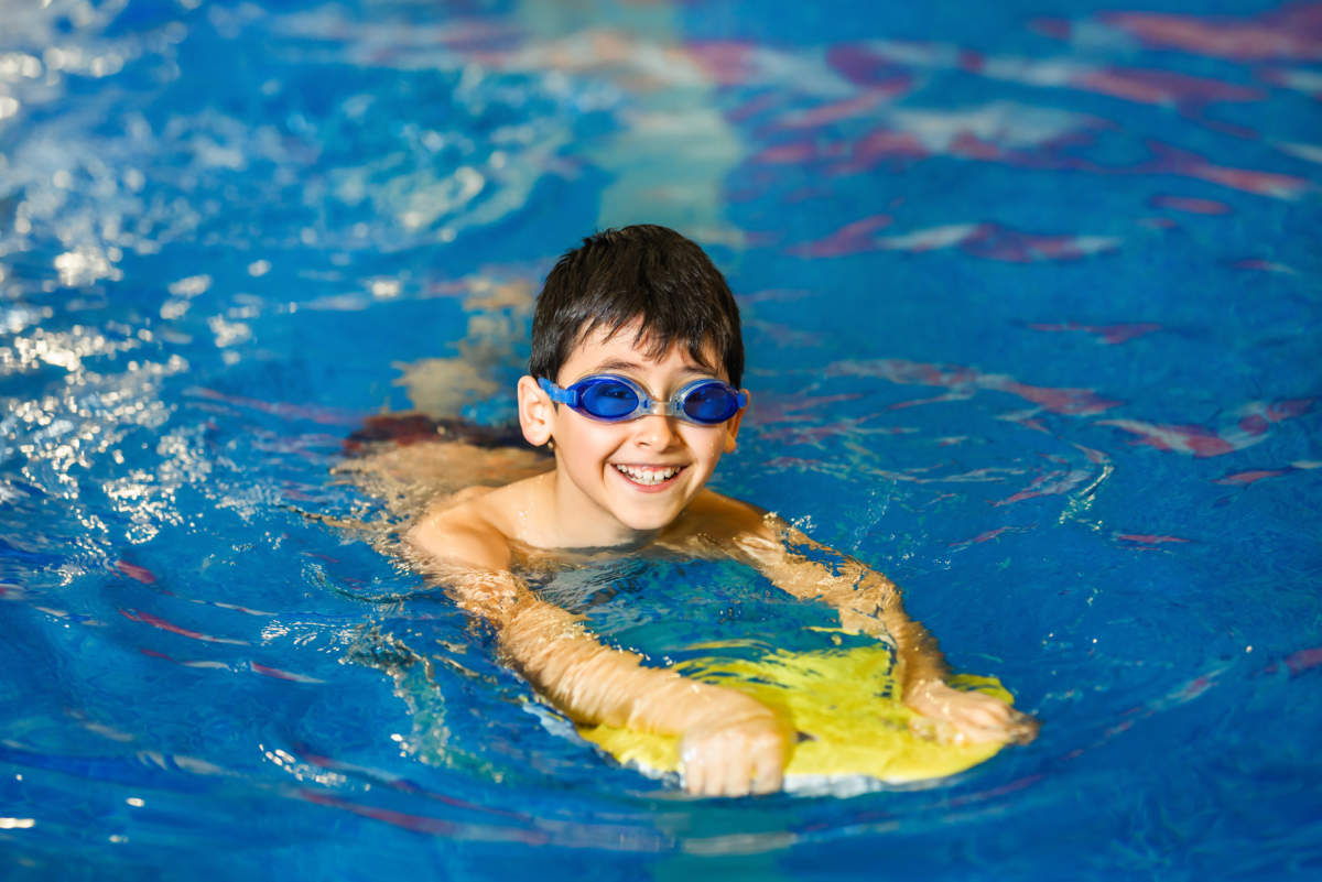 A Little Boy Of 10 Years Learns To Swim In The Sport Pool Using A Board. The Boy Wore Swimming Goggles. The Child Is Cheerful And Happy. Healthy Kid Enjoying Active Lifestyle.