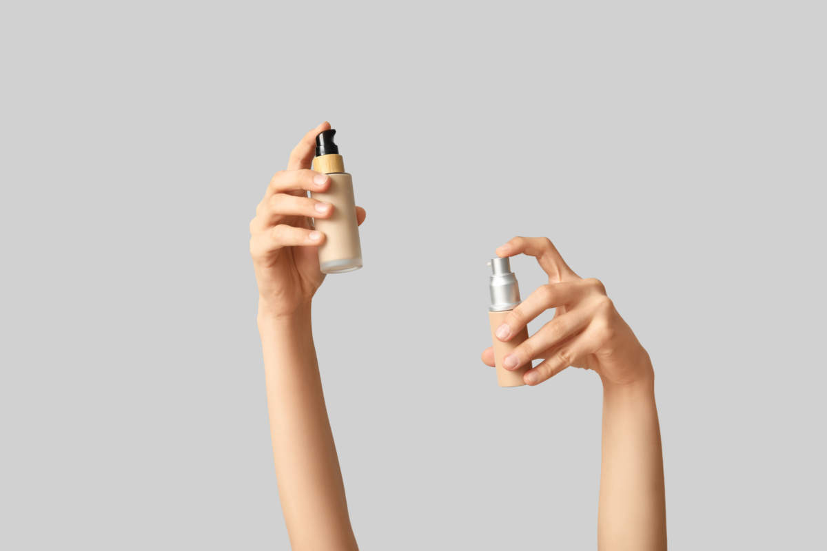 Female Hands With Bottles Of Makeup Foundation On Grey Background