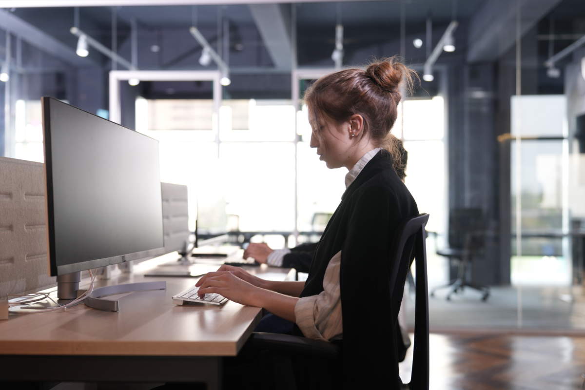Young Businesswoman With An Auburn Bun Works At A Widescreen Desktop Computer Beside Tall Glass Meeting Rooms Flooded With Daylight In The Open Plan Office In A Bright Modern Coworking.